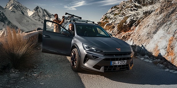 Ein Pärchen ist mit ihrem CUPRA unterwegs. Auf dem Dach befindet sich ein Ski Rack und im Hintergrund sind Schnee bedeckte Berge.