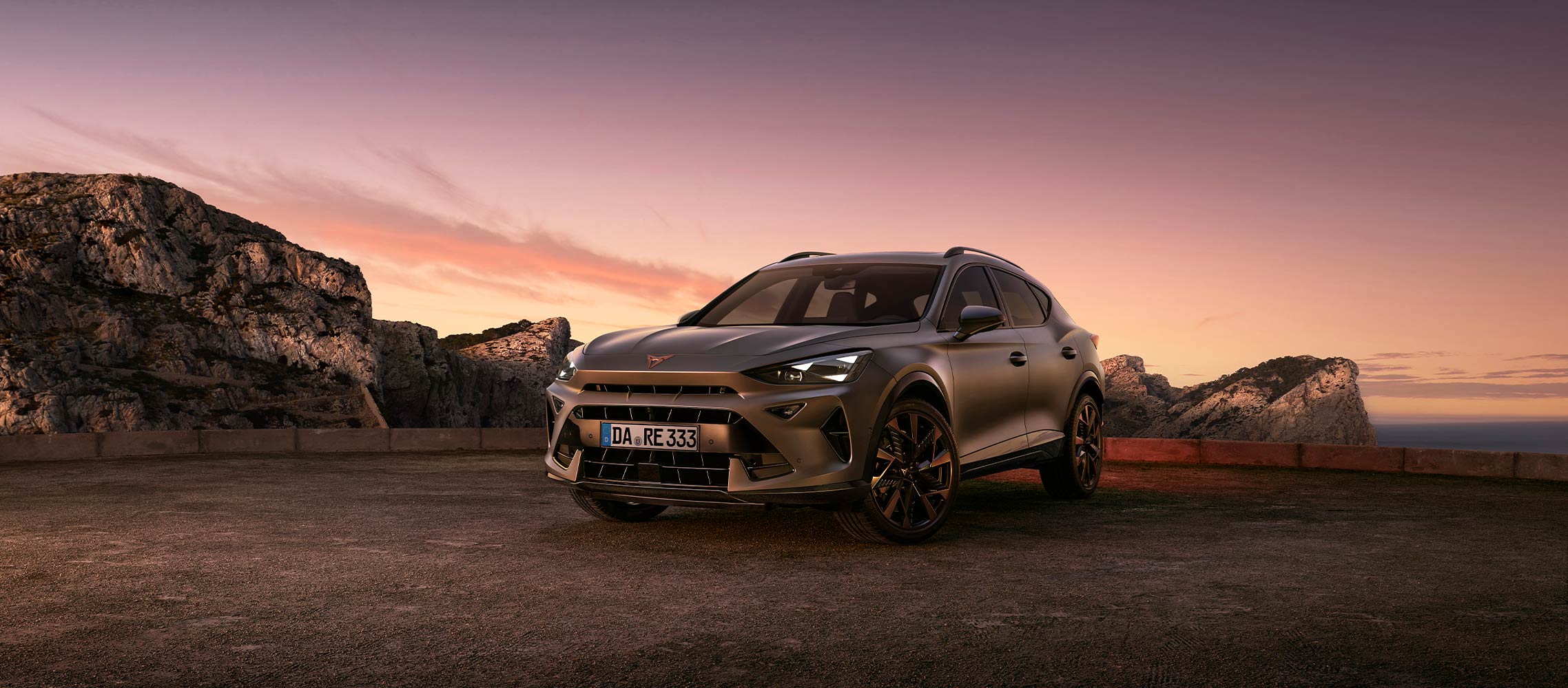 Das Plug-in-Hybrid-Elektrofahrzeug CUPRA Formentor parkt in der Abenddämmerung auf einem Schotterplatz und zeigt eine glatte, metallische Karosserie vor einer Kulisse aus schroffen Felsen und einem himmlischen Sonnenuntergang