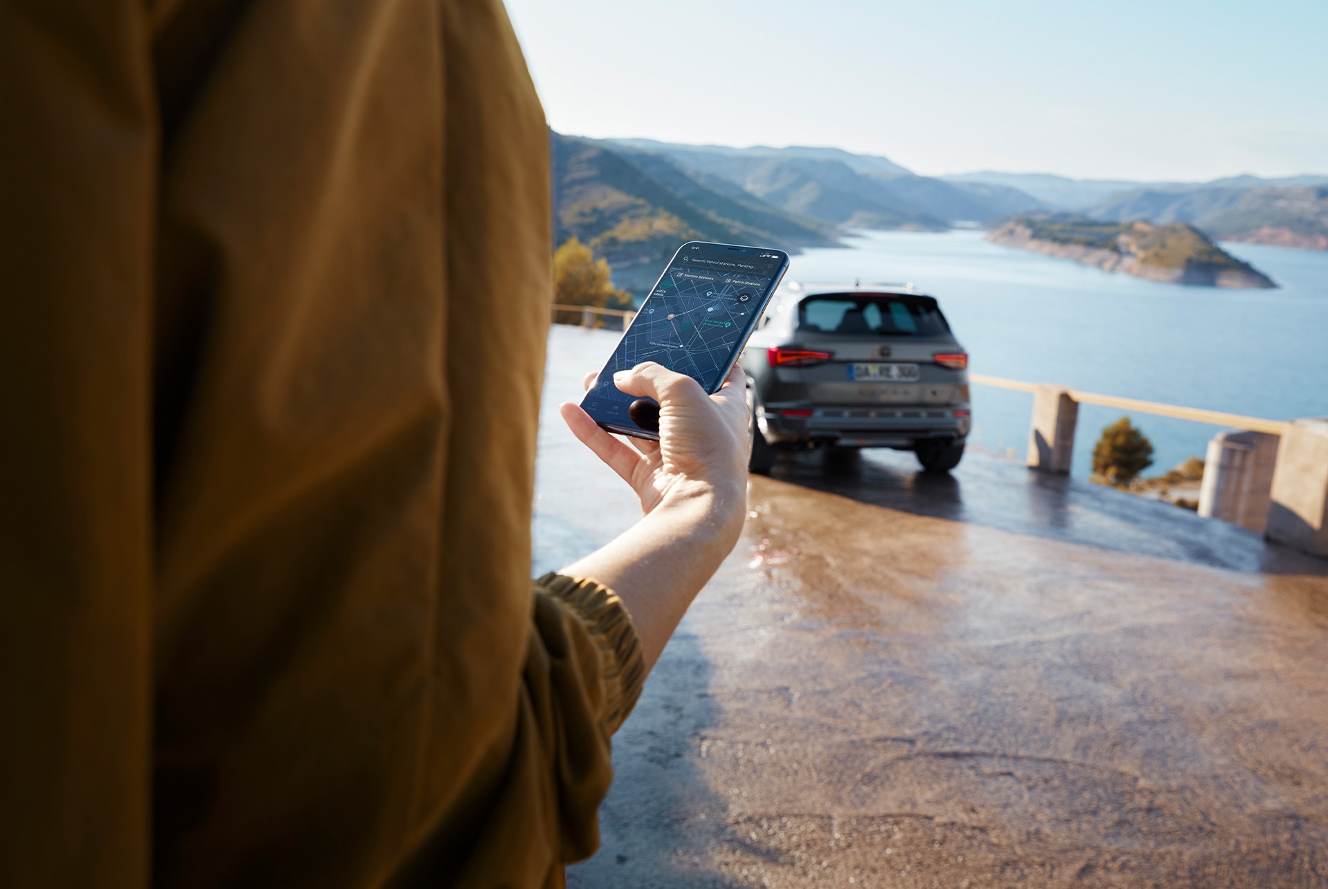 Eine Person hält ein Smartphone, während der CUPRA Ateca im Hintergrund geparkt ist, nahe einer malerischen Aussicht auf Berge und Wasser, und symbolisiert eine vernetzte Welt.