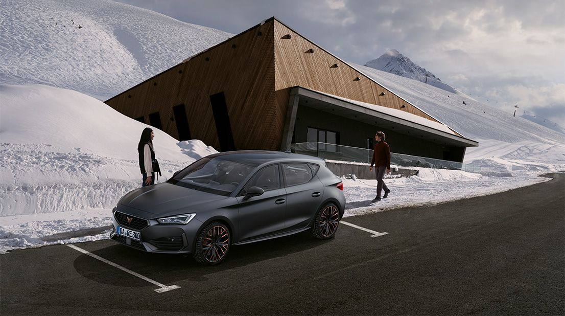 Seitenansicht eines CUPRA Fahrzeugs auf einer freigeräumten Straße, umgeben von Schnee. Eine Frau steht vorne am Auto, während ein Mann von hinten näherkommt.