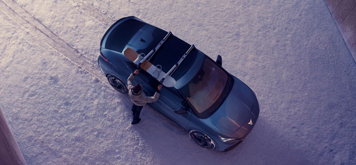 CUPRA Auto von oben mit einem Snowboardhalter, während eine Person das Snowboard anbringt, umgeben von winterlicher Landschaft.